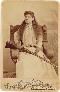 Annie Oakley : Annie Oakley:. Aqui, a lendária atiradora de elite senta-se de forma bastante recatada numa enorme cadeira de estilo renascentista, com uma espingarda no colo. Os seus traços femininos delicados e o cabelo esvoaçante desmentem a sua rudeza a de American Photographer