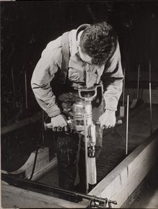 [Um homem utiliza uma máquina manual para trabalhar com um pedaço de madeira].