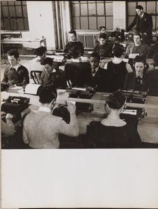 [Pessoas sentadas em frente a máquinas de escrever com auscultadores].