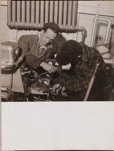 [Dois homens trabalham numa máquina com uma chave de fendas e uma chave inglesa].