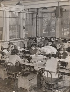 [Mulheres sentadas em secretárias trabalham em máquinas de costura].