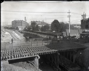 Fotografia horizontal em preto e branco mostrando uma ponte de madeira e trilhos de bonde sobre trilhos de ferrovia parcialmente construídos. Um bonde marcado como Chouteau 2137 está começando a cruzar a ponte na extrema direita. À distância, edifícios de  de Charles Clement Holt