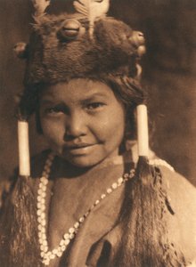 Grande plano de um rapaz índio americano da tribo ou aldeia Klamath. Tem um chapéu de pele com bolotas e penas aplicadas e usa o cabelo em dois rabos de cavalo de Edward Sheriff Curtis