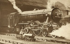 Rapaz a conduzir uma locomotiva a vapor de brinquedo em frente de uma verdadeira (fotografia a preto e branco) de English Photographer