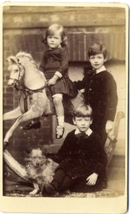 Menina em cima de um cavalo de balanço com um menino de pé e um menino sentado com um cão. de English School