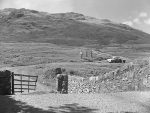 Vista da estrada que passa sobre Hard Knott Pass, duas pessoas de pé perto de um sinal de trânsito e um carro estacionado na berma da relva de Joseph Hardman