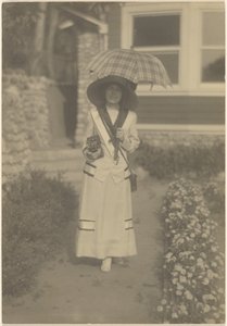 [Mulher em pé no jardim da frente com uma câmara e um guarda-chuva xadrez] de Louis Fleckenstein