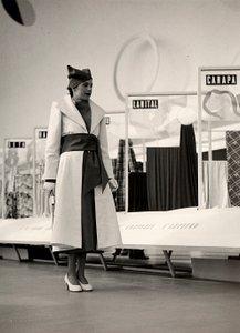 Modelo num sobretudo criado pela casa de alta costura "Lamma". O artigo é recolhido na cintura por um amplo shash, e completado com um chapéu macio. A fotografia foi tirada em um salão utilizado para a exposição de tecidos. (foto a p/b) de Luis Bogino