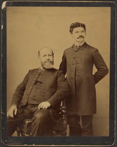 [Homem barbudo não identificado sentado ao lado de um homem com bigode não identificado, de pé]