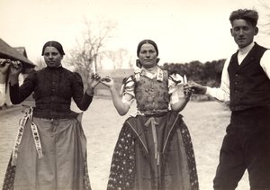 Duas senhoras e um homem, com vestidos tradicionais húngaros, estão de mãos dadas numa dança, segurando algumas velas nas mãos de Rudolf Balogh