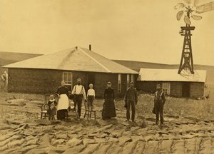 Família de colonos, no exterior da sua casa de relva com um moinho de vento no telhado do edifício adjacente no condado de Custer, Nebraska, c.1885 (fotografia a preto e branco) de Solomon D. Butcher