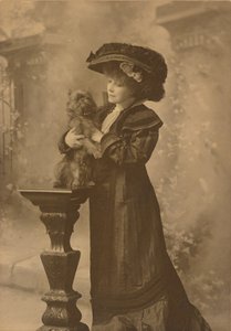 [Sarah Bernhardt de pé, num estúdio, segurando um pequeno cão preto sobre um pedestal de madeira entalhada ornamentado] de Theodore C. Marceau