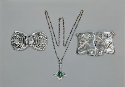 Jóias de prata projetadas para a liberdade. Da esquerda para a direita: Fecho de cintura com design celta entrelaçado, 1904. Conjunto de pingente e corrente com crisoprase de corte cabochão. Fecho de cintura com design repousse em martelado de Archibald Knox