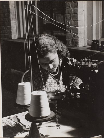 [A mulher utiliza uma máquina de costura com dois carretéis de linha branca]. de Arnold Eagle
