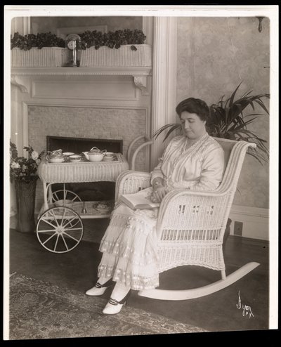 Sra. Howard sentada com um livro em uma cadeira de balanço em sua casa em 200, 5th Avenue, Nova York, 1915-6 (impressão em gelatina de prata) de Byron Company