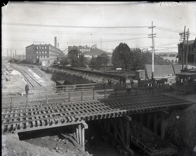 Fotografia horizontal em preto e branco mostrando uma ponte de madeira e trilhos de bonde sobre trilhos de ferrovia parcialmente construídos. Um bonde marcado como Chouteau 2137 está começando a cruzar a ponte na extrema direita. À distância, edifícios de  de Charles Clement Holt