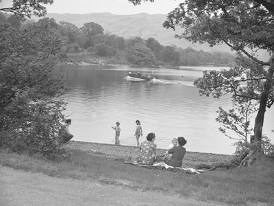 Uma família diverte-se junto a um lago, com um barco no lago e as montanhas ao fundo de Joseph Hardman