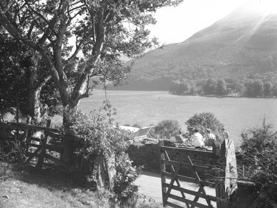 Quatro pessoas sentadas num muro de pedra seca a fazer um piquenique com vista para o lago, o carro estacionado com a bagageira aberta de Joseph Hardman
