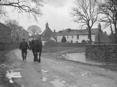 Homem conduzindo um cavalo pela estrada longe da câmara em direção a edifícios de aldeia e igreja; arquitetura; religioso; eclesiástico; de Joseph Hardman