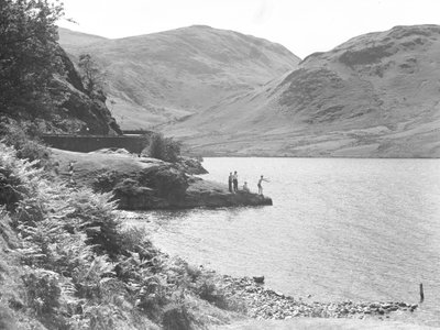 Três homens e uma mulher num afloramento rochoso no lago a pescar, com as montanhas em fundo de Joseph Hardman