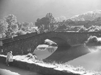 Mulher sentada numa parede a estudar um mapa com um rio e uma ponte de pedra ao fundo de Joseph Hardman