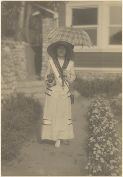 [Mulher em pé no jardim da frente com uma câmara e um guarda-chuva xadrez] de Louis Fleckenstein