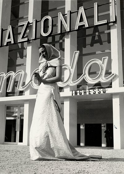 Modelo em vestido de noiva criado pela casa de alta costura "Lamma". O artigo caracteriza-se por um toucado de estilo monástico, segurado no pescoço por um alfinete brilhante na forma de um cacho de uvas. (foto a p/b) de Luis Bogino