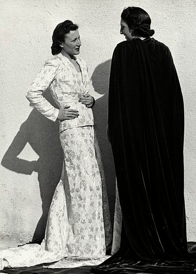 Dois modelos apresentam criações da casa de alta costura italiana "Dragoni". Um modelo veste uma roupa em material fralda composta por uma jaqueta e saia comprida com trem. O outro modelo, visto de trás, veste um longo manto de veludo. (foto a p/b) de Luis Bogino