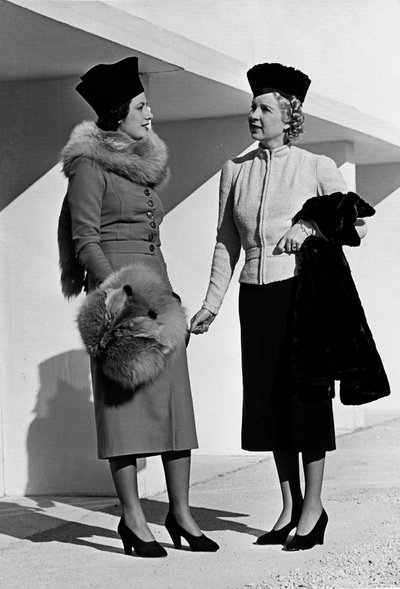 Dois modelos apresentam artigos de vestuário de dia criados pela casa de costura "Fumach". A modelo à esquerda usa um vestido complementado por estola de raposa e muff. A modelo à direita veste um fato composto por um casaco curto e uma saia. (foto a p/b) de Luis Bogino