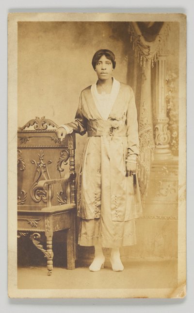 Postal fotográfico de uma mulher ao lado de uma cadeira de madeira, 1904-1918. de Unbekannt