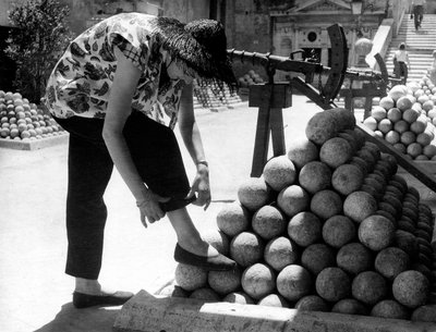 Uma mulher ajusta o punho das calças enquanto apoia o pé numa pilha de balas de canhão no recinto do Castelo de Santo Ângelo, em Roma. de Unknown photographer
