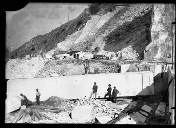 Três homens observam o trabalho de várias escavadoras nos Alpes Apuanos. Atrás delas há um enorme bloco de mármore já quadrado e pronto para ser transportado para o vale (foto a p/b)