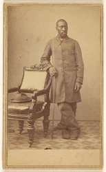[Soldado negro de pé junto a uma cadeira, com chapéu no assento]