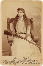 Annie Oakley : Annie Oakley:. Aqui, a lendária atiradora de elite senta-se de forma bastante recatada numa enorme cadeira de estilo renascentista, com uma espingarda no colo. Os seus traços femininos delicados e o cabelo esvoaçante desmentem a sua rudeza a