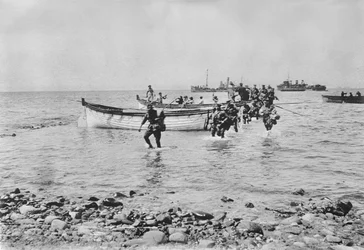 Soldados australianos atravessando a água ao desembarcarem em Anzac Cove, Gallipoli, em 25 de abril de 1915, após serem transportados em barcos de desembarque de navios maiores ao fundo, em 25 de abril de 1915 (