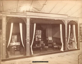 Exposição de James Shoolbred & Co. -- Edifício Principal, Três quartos com móveis. A sala à esquerda contém três armários e um lavatório, uma torre e um espelho; a sala central contém uma mesa, cadeiras, um sofá e um aparador com um espelho e uma escultura