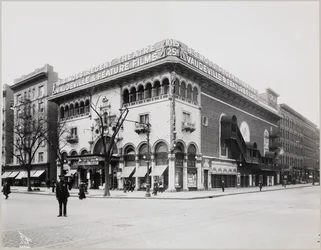 Um enorme teatro com uma fachada elaborada, completa com arcos mouriscos e varandas. Um grande letreiro no topo do edifício diz „B.S. Moss Regent Theatre Vaudeville & Feature Films“. Um cartaz a anunciar Olga Petrova em „The Heart of a Painted Woman“ está