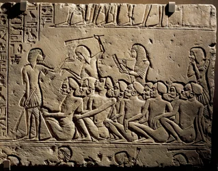 Um grupo de prisioneiros negros rodeados por guardas armados com mocas, enquanto um escriba redige um relatório, relevo do túmulo de Horemheb em Sacará