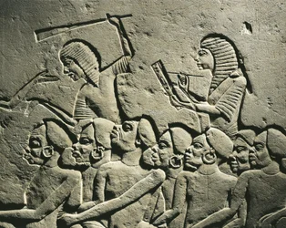 Um grupo de prisioneiros negros rodeados por guardas armados com mocas, enquanto um escriba redige um relatório. Relevo do túmulo de Horemheb em Sacará, pormenor