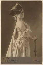 [Mulher não identificada, de chapéu escuro, de pé, com as mãos apoiadas numa bengala].