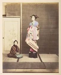 [Geisha,] Retrato de estúdio de pé de uma jovem mulher em sapatos elevados, o seu jovem atendente ajoelhado no chão ao seu lado, Fotógrafo: Colecção Lucas desconhecida: Registo fotográfico da digressão mundial de Charles James e Morton P, Luc