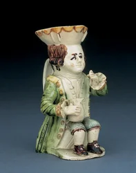 Jarro Toby do beberrão, o chapéu com bordo recortado castanho e amarelo, o cabelo castanho inciso preso em rabicho com uma roseta branca, sentado com um jarro em forma de balaústre sobre o joelho, c. 1770