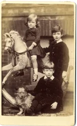 Menina em cima de um cavalo de balanço com um menino de pé e um menino sentado com um cão.