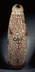 Escudo oval Asmat, esculpido em relevo com ornamento em espiral incorporando figuras altamente estilizadas, borlas entrançadas até a borda (madeira, ráfia e penas)