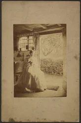 [Mulher de vestido comprido às riscas sentada no interior da sala].