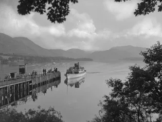 Um barco que transporta pessoas num lago, perto de um cais, com as montanhas ao fundo