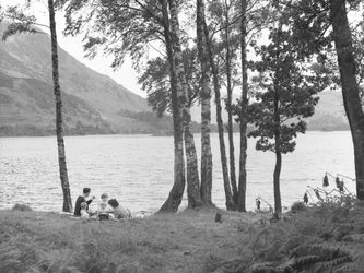 Uma família a descontrair junto ao lago, com árvores à direita e montanhas ao fundo