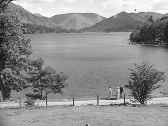 Um homem, uma senhora e possivelmente a sua filha passeiam ao longo da margem de um lago, com as montanhas em fundo