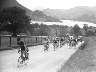 Grupo de ciclistas a subir uma estrada seguido de carros, com o lago e as montanhas em fundo