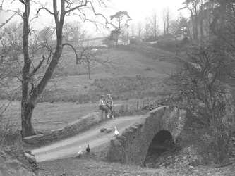Duas raparigas sentadas numa ponte de pedra sobre o rio Cocker, no sopé de Scale Hill, observando gansos no caminho em Loweswater